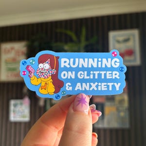 May include: A blue sticker with the text "RUNNING ON GLITTER & ANXIETY" features a cartoon character in a yellow sweater holding a pink toy gun. The character has brown hair and a polka-dotted face. The sticker has a cloud-like border.