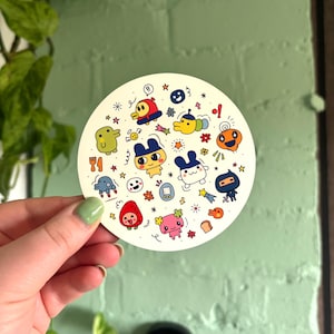 May include: A round sticker with a white background and colourful cartoon characters. The characters include a duck, a ninja, a bunny, a strawberry, and a caterpillar. There are also stars, flowers, and other small details.
