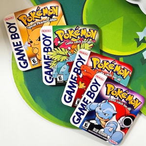 May include: Four vintage Game Boy Pokemon game cartridges. The cartridges are blue, red, green, and yellow. Each cartridge features a different Pokemon character and the text "Gotta catch 'em all!"