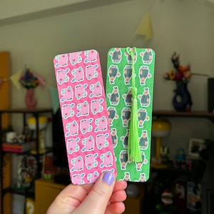 May include: Two bookmarks with different designs. One bookmark is pink with a repeating pattern of pink pigs with green details. The other bookmark is green with a repeating pattern of green stick figures wearing green hats. Both bookmarks have green tassels.