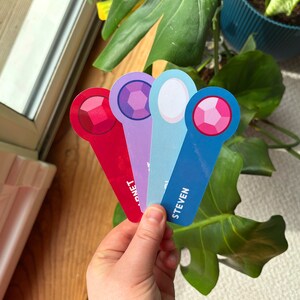 May include: Four colourful bookmarks with gem-shaped designs and text. The bookmarks are red, purple, blue, and white. The text on the bookmarks reads "Steven", "Connie", "Pearl", and " Garnet".