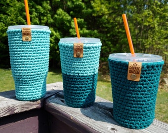 May include: Three crocheted coffee cup sleeves in teal and turquoise colours. The sleeves have different designs and text, including "Shhh... Coffee", "Coffee & Cats", and "Coffee Queen".