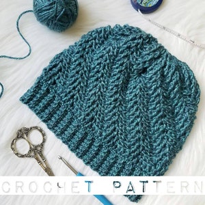 May include: Teal blue crochet beanie hat with a chevron stitch pattern. The hat is laying on a white fluffy surface with a crochet hook and scissors nearby.
