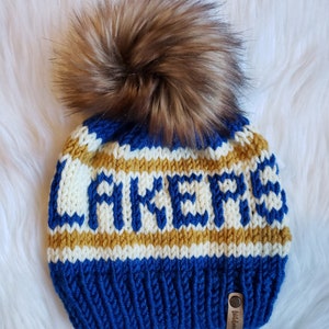 May include: Blue knitted beanie with a white and gold "LAKERS" design and a brown faux fur pom pom. The beanie has a brown leather tag with the word "Wildwood" on it.