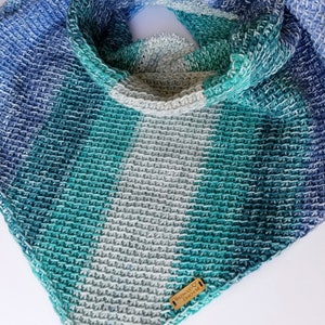 May include: A crocheted infinity scarf in shades of blue, green, and grey. The scarf has a textured stitch pattern and features a small brown leather tag with the text "Wildwood Designs ME".