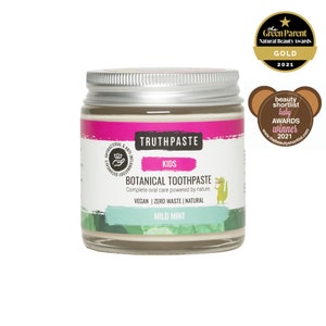 Op de afbeelding: Een glazen pot Truthpaste Kids Botanical Tandpasta met een zilveren deksel. Het etiket toont de merknaam, productdetails en een groene krokodil-illustratie. Er zijn onderscheidingen zichtbaar. De tandpasta heeft een milde muntsmaak.