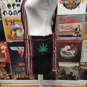 May include: A black and green striped crossbody bag with a green marijuana leaf design. The bag has a long strap and a flap closure.