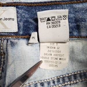 May include: A close-up of a light blue denim jean label with white text. The label reads "Size 3", "Inseam 32", "ZJ23U1508", "Calvin Bootcut", "Double Stone Wash", "5503053-32-18", and "000145".