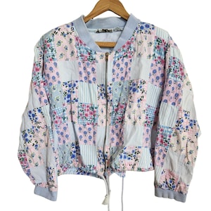 May include: A patchwork bomber jacket with a floral pattern in pink, blue, and white. The jacket has a zip-up front, a drawstring hem, and light blue ribbed cuffs and collar. The fabric is a mix of floral and striped patterns.
