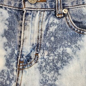 May include: Close-up of a pair of light blue denim jeans with a white bleach splatter pattern. The jeans have a button fly and a metal button with the letter "G" on it.