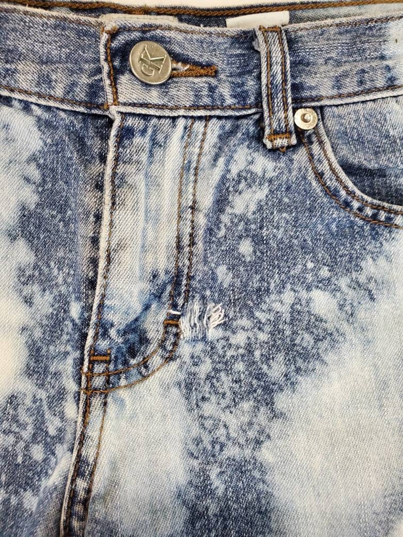 May include: Close-up of a pair of light blue denim jeans with a white bleach splatter pattern. The jeans have a button fly and a metal button with the letter "G" on it.