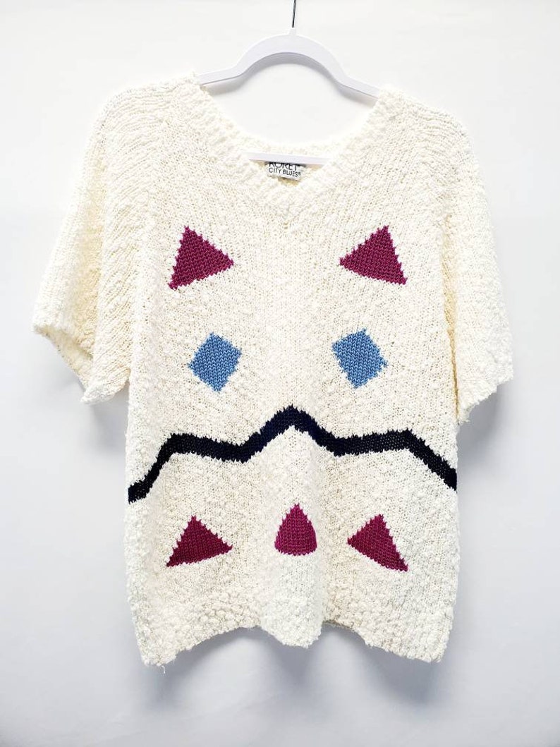 May include: A white knitted sweater with a geometric pattern in shades of pink, blue, and black. The sweater has short sleeves and a v-neck. The label reads "Robert City Blues".
