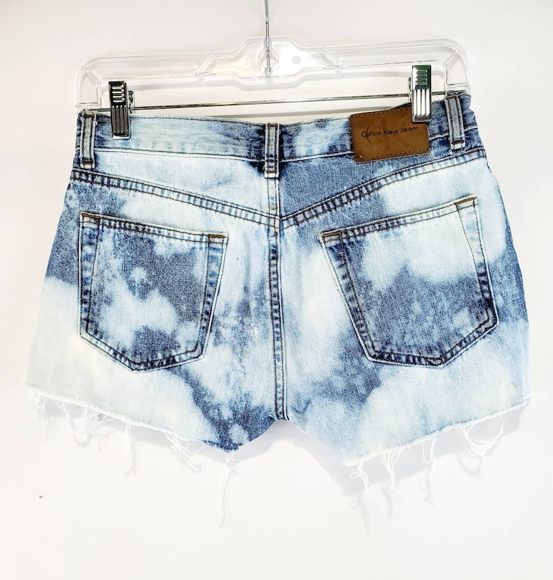 May include: Light blue denim shorts with a distressed hem and a brown leather patch that says "Calvin Klein Jeans".