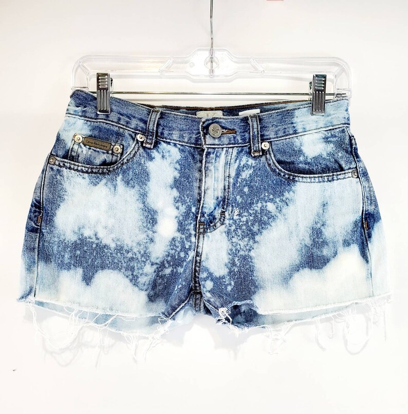May include: Light blue denim shorts with a bleach-splattered design. The shorts have a frayed hem and a button fly.
