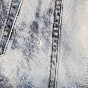 May include: Close-up of light blue denim fabric with a single seam running vertically down the center. The fabric has a faded, distressed look.