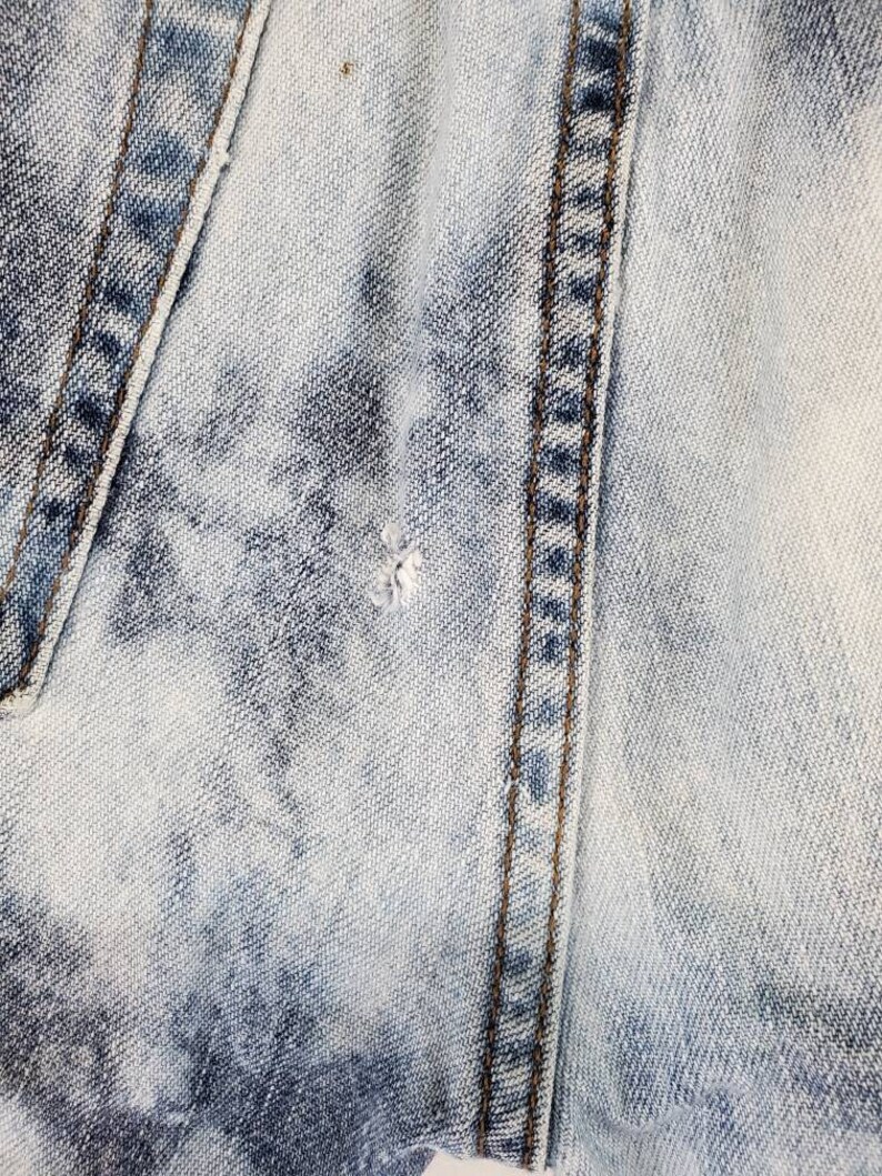 May include: Close-up of light blue denim fabric with a single seam running vertically down the center. The fabric has a faded, distressed look.
