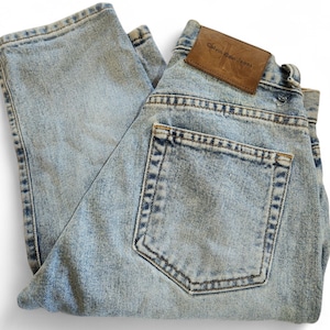 May include: Light blue denim jeans with a classic five-pocket design. The jeans feature a faded wash and a brown leather patch with the "Calvin Klein Jeans" logo. The jeans are folded, showcasing the back pocket and the brand label.