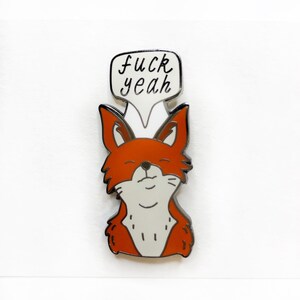 May include: A pin featuring a cartoon fox with orange fur, a white belly, and a speech bubble that says "fuck yeah". The fox has a relaxed expression with closed eyes. The pin has a silver outline.