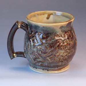 May include: A handcrafted ceramic mug with a dark brown and teal glaze. The mug features embossed floral and geometric patterns. The handle has gold accents. The interior is a light tan color.