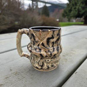 May include: A handmade ceramic mug with a unique textured design. The mug features a beige and black pattern with a dark interior and a curved handle. The mug is approximately 4 inches tall.