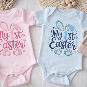 May include: Two baby onesies, one pink and one light blue, with the text "My 1st Easter". Each onesie has bunny ears and paw prints. A beige beanie and wooden beads are also in the image.