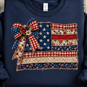 May include: A navy blue long-sleeve sweatshirt featuring a central graphic of a patchwork American flag. The flag design incorporates various fabric patterns including red and white gingham, floral prints, and lace, topped with a large decorative bow.