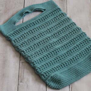 May include: A teal-colored, rectangular crochet tote bag with a single handle. The bag features a textured, ribbed pattern throughout its body. The handle is also crocheted and is attached to the top of the bag. The bag is lying on a light-colored wooden surface.