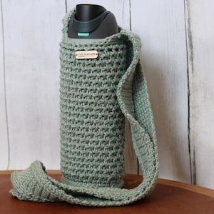 May include: A sage green crochet water bottle holder with a matching strap. The holder has a small wooden tag that reads "rich textures". The bottle is black with a teal accent. The holder is designed to fit a standard water bottle.
