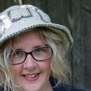 May include: A woman with blonde hair wearing a white crocheted hat with gray accents and glasses. She is smiling and looking at the camera.