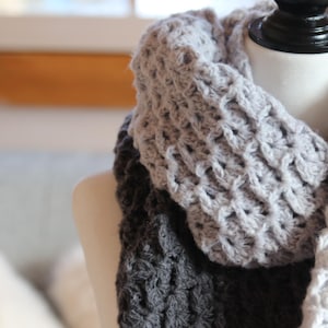 Puede incluir: Bufanda de crochet hecha a mano en tonos grises y blancos, drapeada sobre un maniquí. La bufanda presenta un patrón de puntada texturizada y está diseñada para usarse alrededor del cuello. El degradado de color añade interés visual al accesorio.