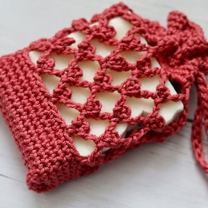 Puede incluir: Una bolsa de jabón de ganchillo de color rosa polvoriento con una barra de jabón blanca en su interior. La bolsa tiene un cierre de cordón.