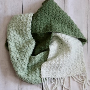 May include: A handmade crochet scarf in shades of green and white. The scarf is folded, showcasing the different colors and textures of the yarn. The scarf has a fringed edge on one end.