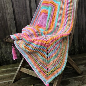 May include: A colourful crocheted throw blanket with pink, orange, green and blue stripes. The blanket is draped over a wooden chair with tassels hanging from the bottom corners.