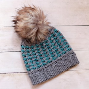 May include: A crocheted beanie featuring a ribbed grey brim and a textured body with alternating rows of grey and teal yarn. The hat is topped with a large, fluffy faux fur pom-pom in shades of brown, tan, and cream, adding height and texture.