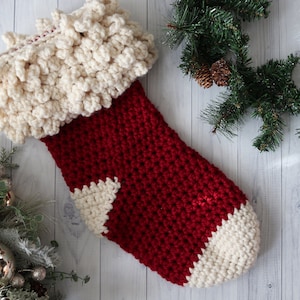 Peut inclure: Une chaussette de Noël rouge et blanche en crochet avec un revers blanc et moelleux. La chaussette est accrochée à un fond en bois blanc.