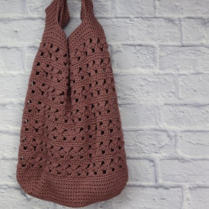 May include: A dusty rose crochet tote bag with a floral pattern. The bag has a rounded bottom and two shoulder straps. The bag is hanging against a white brick wall.