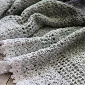 Pode incluir: Um cobertor feito à mão, de crochê, em tons de cinza e branco. O cobertor apresenta um padrão de ponto texturizado e uma borda ondulada. A cor transita do branco para o cinza, criando um efeito gradiente. O cobertor é macio e parece ser feito de um fio leve.
