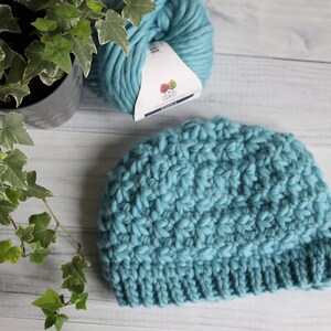 May include: A teal blue crocheted beanie hat made with chunky yarn. The hat is lying on a white wooden surface next to a ball of teal blue yarn.