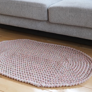 May include: An oval, hand-crocheted rug in pink, grey, and white hues. The rug is positioned on a wooden floor, partially beneath a grey sofa. The texture of the rug is evident, highlighting the individual stitches.