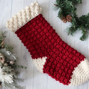Puede incluir: Un calcetín de Navidad de crochet rojo y blanco con un puño blanco. El calcetín está colgado en un fondo de madera blanco.