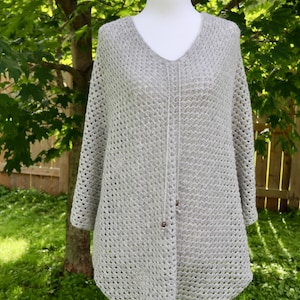 May include: A light gray crochet poncho featuring a V-neckline and an openwork diamond pattern. The garment includes a central drawstring cord finished with small, dark, spherical beads at the ends, hanging down the front center.