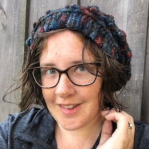 May include: A woman with short, wet hair wearing a blue, red, and black knit beanie and brown-rimmed glasses. She is smiling and looking at the camera.