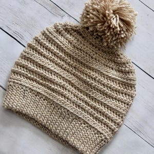 May include: A beige crocheted beanie with a fluffy pom pom on top. The beanie has a ribbed texture and is made with a soft yarn.
