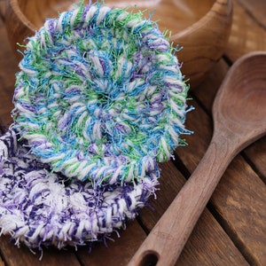 May include: Two crocheted dishcloths, one in shades of blue, green, and white, and the other in shades of purple, white, and blue, on a wooden surface.