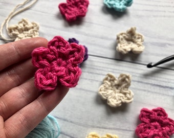 May include: A close-up of a pink crocheted flower. The flower is being held in a hand. There are other crocheted flowers in the background, including white, blue, and purple.