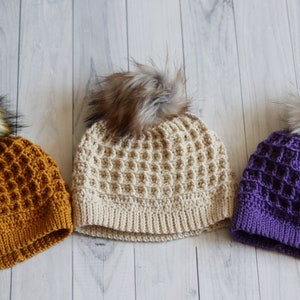 May include: Three crocheted hats with faux fur pom poms. The hats are in mustard yellow, cream, and purple.