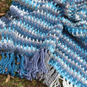 May include: A crocheted throw blanket with a striped pattern in shades of blue, gray, and white. The blanket has fringe along the bottom edge.