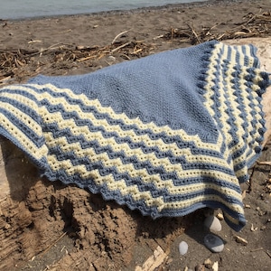 May include: A blue and white crocheted shawl with a chevron pattern on the border. The shawl is draped over a piece of driftwood on a sandy beach.