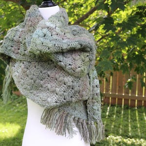 May include: A hand-crocheted shawl featuring a textured cluster stitch pattern in variegated shades of sage green, olive, and soft brown. The triangular garment is draped over a mannequin and finished with long, matching fringe along the bottom edge.