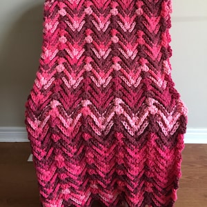 May include: A pink and red chevron crochet throw blanket with tassels. The blanket is made with a textured stitch and has a soft, cozy feel.